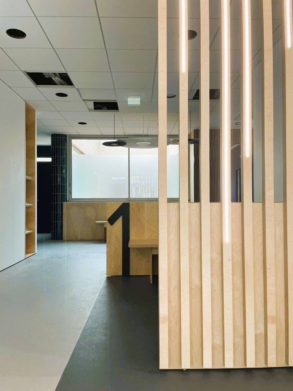 La claustra en tasseaux de bois et ses lignes de lumière, la salle 1 derrière